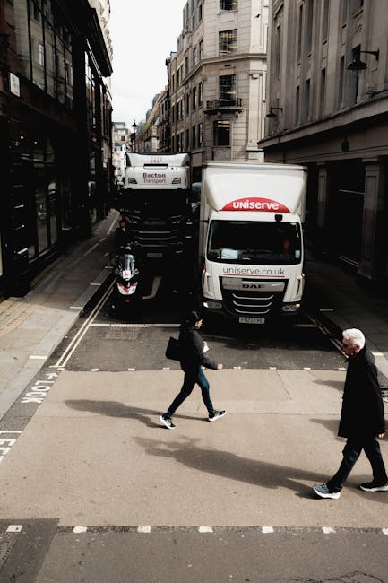 A busy urban street scene viewed from an elevated perspective showing a small pedestrian crossing with two people walking across it—one woman dressed in dark clothing and carrying a bag, and an older man with white hair also walking. Behind them, two moving trucks with branding from Uniserve and Becton Transport are parked on the road, with the back of one truck extending onto the pavement. A scooter is parked near the curb. The street is lined with tall, historic buildings featuring fire escapes and large windows, with a narrow section of the road reserved for loading and unloading. Overcast lighting illuminates the scene, capturing elements typical of home relocation and furniture transport logistics, with visible boxes, packing materials, and equipment associated with house removals, all within a busy city environment. Kennington Removals provides these services, ensuring efficient and professional moving processes in the area.