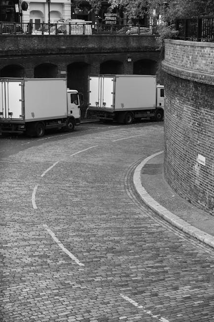 A black-and-white photograph of a cobbled residential street featuring three white moving trucks parked along the curb, arranged in a line under an arched brick bridge. The trucks are positioned to load or unload household furniture and boxes, which are not visible in the image. The street has a gentle curve, with a brick wall on the right side and a metal fence on top of the bridge, beyond which trees and residential buildings can be seen. The scene captures the logistics involved in home relocation, with the trucks ready for furniture transport, and a quiet, orderly environment typical of a planned house removal. The image emphasizes the loading process, the use of a suitable vehicle for moving services, and the street environment where furniture packaging materials and possibly blankets or straps might be used during the loading process, supporting the relocation activities carried out by Kennington Removals.