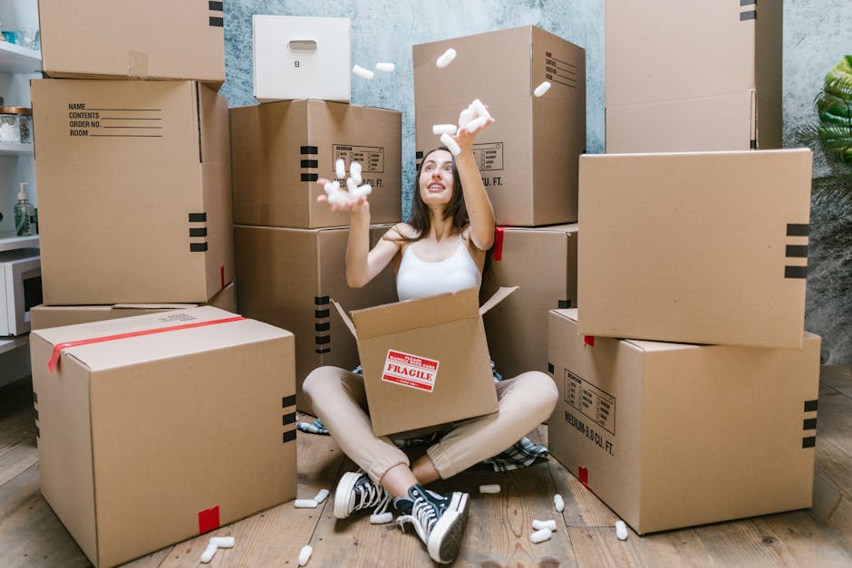 A young woman sitting cross-legged on a wooden floor inside a room, surrounded by numerous packed cardboard moving boxes in various sizes, some with red 'Fragile' labels and others with black printed markings. She is wearing casual clothing, including a white tank top and beige pants, and has a cheerful expression while tossing white packing peanuts into the air. The boxes are stacked behind her, filling most of the space, and a doorway is visible nearby, suggesting she is in the process of packing or preparing for a home relocation. The room has a textured, light-colored wall, and a small green plant can be seen in the corner. This scene illustrates the packing and loading process associated with house removals, typically handled by professional services such as Kennington Removals, ensuring safe furniture transport and careful handling of belongings during moving projects.