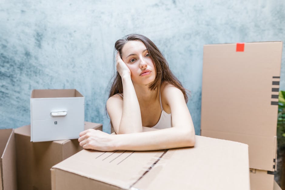 A young woman sitting cross-legged on a wooden floor inside a room, surrounded by numerous packed cardboard moving boxes in various sizes, some with red 'Fragile' labels and others with black printed markings. She is wearing casual clothing, including a white tank top and beige pants, and has a cheerful expression while tossing white packing peanuts into the air. The boxes are stacked behind her, filling most of the space, and a doorway is visible nearby, suggesting she is in the process of packing or preparing for a home relocation. The room has a textured, light-colored wall, and a small green plant can be seen in the corner. This scene illustrates the packing and loading process associated with house removals, typically handled by professional services such as Kennington Removals, ensuring safe furniture transport and careful handling of belongings during moving projects.