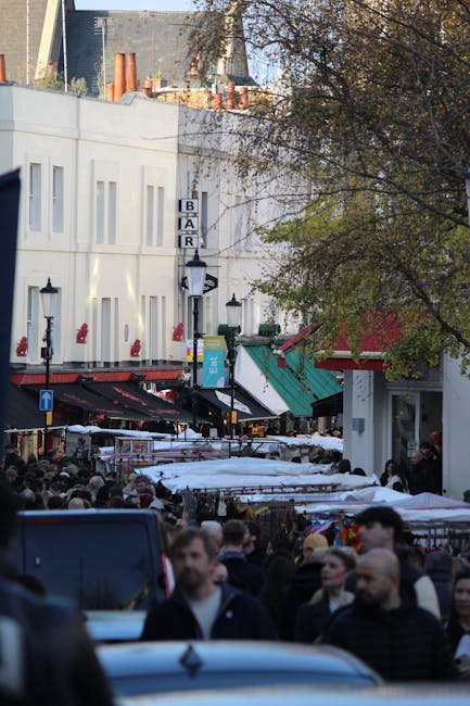 A densely crowded street scene with numerous pedestrians walking along the pavement outside shops and a bar, under a clear sky. The street features outdoor market stalls covered with white and beige f