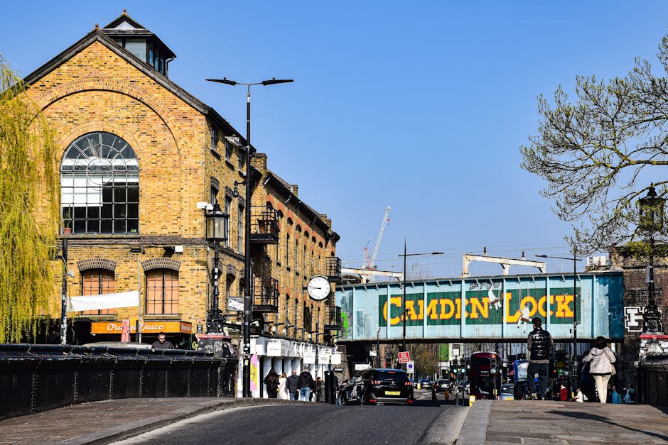 A densely crowded street scene with numerous pedestrians walking along the pavement outside shops and a bar, under a clear sky. The street features outdoor market stalls covered with white and beige f