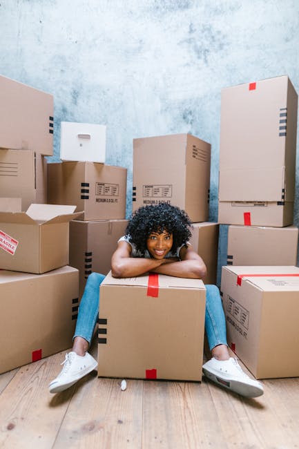 A young woman with curly black hair, wearing a white top, blue jeans, and white sneakers, is sitting on a light wooden floor surrounded by numerous brown cardboard moving boxes of various sizes, some with black markings and red labels. She is smiling and resting her chin on her arms, which are folded over a large box in front of her. The room has a textured light blue wall and natural light coming from an unseen source. The boxes are stacked in the background and foreground, indicating an active packing or home relocation process. This scene illustrates the packing stage of house removals by Kennington Removals, highlighting their professional packing and moving services, including furniture and household item transport during a home relocation or same-day emergency move.