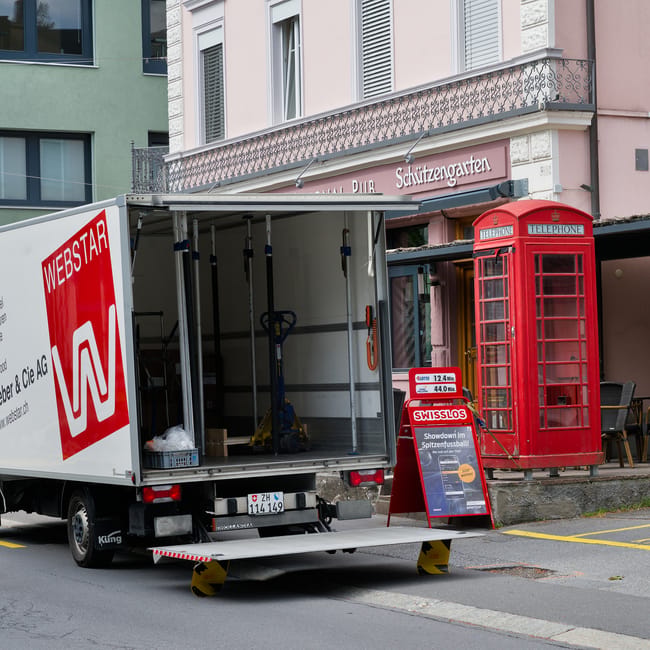 A moving truck with an open loading area parked on a street in front of a building with a balcony and signage. Inside the truck, visible packing and moving equipment includes a hand truck, a blue adjustable strap, and some plastic bags. The truck is positioned at a slight incline with wheel chocks for stability. Nearby, a classic red telephone booth stands on the pavement next to a freestanding sandwich board sign advertising Swiss cheese. The building behind features pastel pink and white exterior walls with window shutters, and a sign indicating a pub with a beer garden beneath the balcony. This scene depicts a home relocation process involving furniture transport and packing activities, with the logistics of loading household goods into the vehicle for a house move. The environment is well-lit with natural daylight, and overall, it captures a typical street scene during a household removal service provided by Kennington Removals.