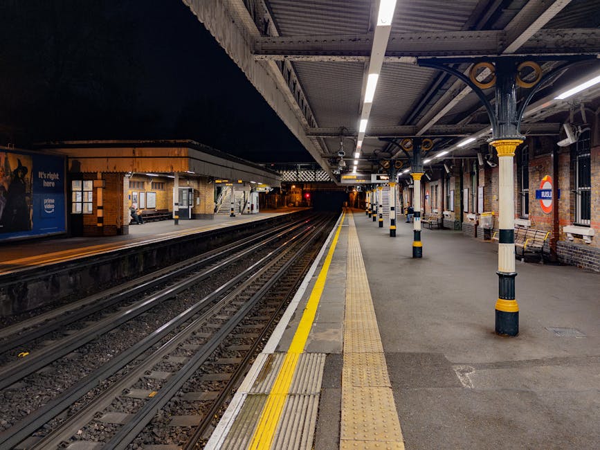 At Kennington Station, the underground platform is illuminated by overhead lighting, showing parallel train tracks with gravel beds and a yellow safety line along the edge. The platform is sheltered by a metal canopy supported by decorative columns painted with black and yellow bands, providing shelter for passengers. On the far side of the platform, a small brick building houses ticket machines and information boards, with a person seated near the entrance. Benches are placed along the platform for waiting passengers, and the station features brick walls with large windows and advertising posters. This setting is relevant to house removals and relocation logistics, as it highlights the surrounding environment where moving vans might access the station for furniture transport or loading operations, with the platform prepared for the arrival or departure of luggage and moving boxes during a home relocation process facilitated by [COMPANY_NAME].