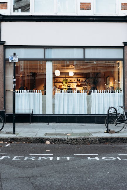 Photograph of the exterior of a shop or cafe with large glass windows and white curtains partially drawn inside, illuminated by warm lighting. The storefront is located on a street with a sidewalk, where two bicycles are parked and secured to bike racks on either side of the entrance. The pavement features a marked parking bay with the words 'PERMIT ONLY' painted in white, indicating parking restrictions. The building has a two-tone façade with a black lower section and a brick upper section. Inside, shelves with various items and interior lighting are visible through the windows. The scene captures a quiet moment during daytime, with no visible people or moving activity, but it relates to urban environment considerations relevant to house removals and packing logistics.