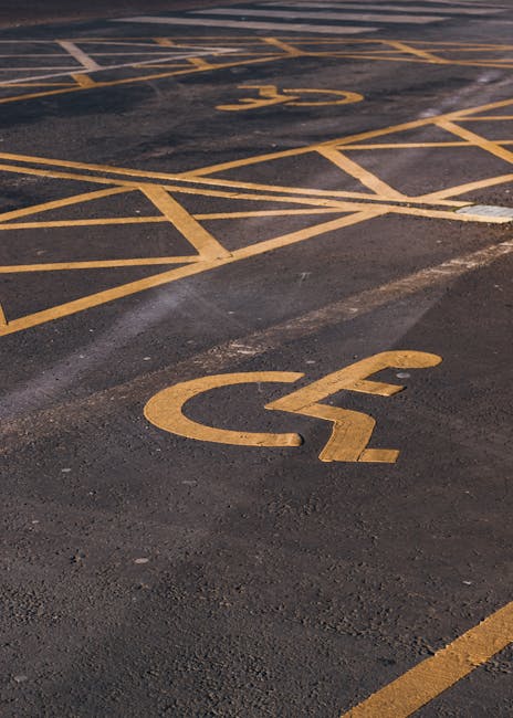 A close-up view of a designated disabled parking space in an outdoor parking lot, featuring yellow-painted wheelchair symbols on dark asphalt. The space is marked with clearly visible yellow lines and symbols, with some overlapping lines indicating adjacent parking bays. The surface shows signs of wear and slight discoloration around the markings. The parking area is part of a commercial or public space, which may be used for vehicle loading and unloading during house removals or home relocation processes. The image emphasizes the importance of accessible parking facilities for moving services like those provided by Kennington Removals, especially when transporting furniture, boxes, and appliances to or from a property in Lambeth or surrounding areas.