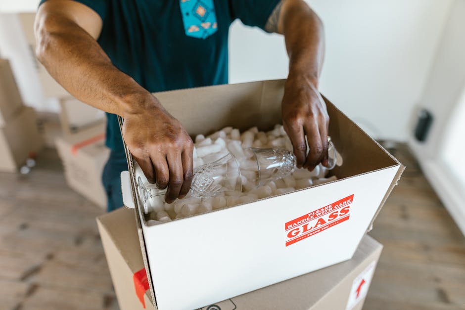 A person’s hands are shown placing a clear glass bottle into a cardboard box filled with white packing foam for protection. The individual is wearing a blue shirt, and the scene is set indoors, on top of a white surface, with a wooden floor visible in the background. The box has a red and white label reading 'GLASS' to indicate it contains fragile items, and it is positioned on a small platform or table. This image captures the careful packing process involved in home relocation and furniture transport, emphasizing the importance of proper packing materials and careful handling during moving and removals, as carried out by Kennington Removals. The lighting is natural, highlighting the focus on secure packing and the protective measures used to safeguard fragile household items during the loading process.