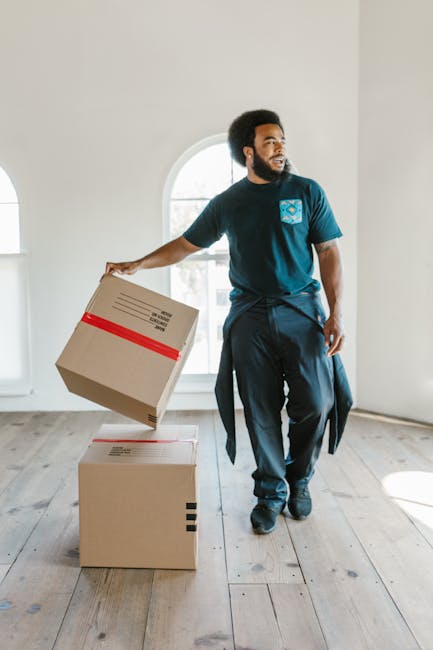 A man with dark curly hair, a beard, and medium skin tone is inside a bright, empty room with white walls and wooden flooring, participating in a home relocation process. He is wearing a navy blue t-shirt with a logo on the chest, dark trousers, and has a piece of clothing tied around his waist. The man is holding a medium-sized cardboard box, which appears to be sealed with red tape and has shipping labels on top. Two additional cardboard boxes are positioned on the floor in front of him, one stacked directly on top of the other, indicating packing and moving activities. The room features a large, arched window allowing natural light to flood the space, creating a well-lit environment. Visible in the background are no furnishings or decorations, emphasizing the ongoing packing or furniture transport process. The scene aligns with the services provided by Kennington Removals for house removals and furniture transport, observed through the careful handling of boxes and the empty interior ready for relocation activities.