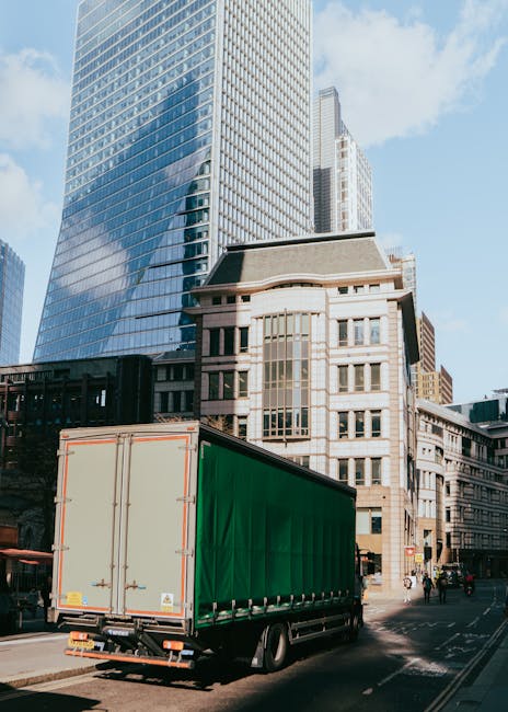 A large green cargo truck parked on a city street during daytime, with tall modern office buildings and historical architecture visible in the background. The truck is positioned near the curb, ready 
