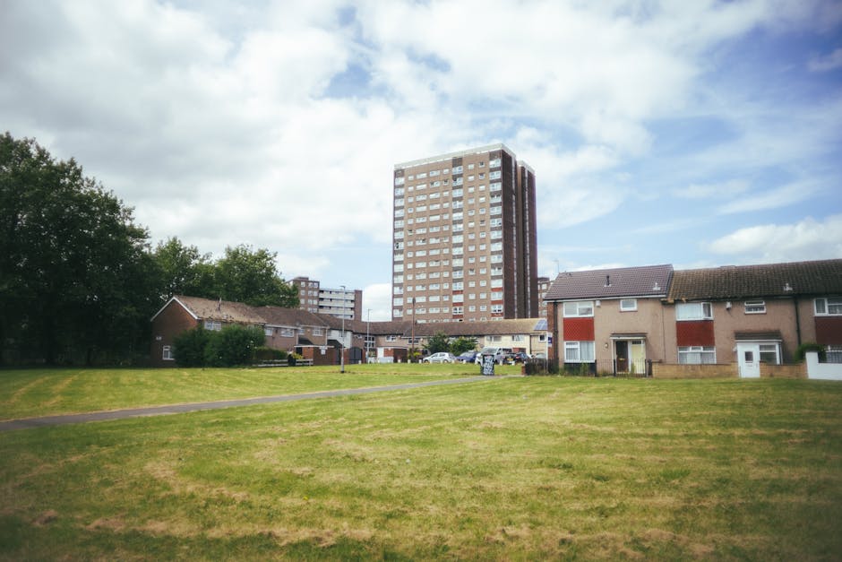 A view of a residential area showing a large high-rise apartment building in the background, with a cluster of single-story houses featuring pitched roofs and small front gardens in the foreground. The houses are made of brick and have white-framed windows, with some displaying additional red and white paneling. A paved pathway runs across a grassy lawn in front of the houses, leading toward the apartment building. On the right side, there are a few cars parked near the houses, and a pickup truck is parked on the street closer to the high-rise. The image appears to be taken on a partly cloudy day with a mix of sun and clouds, and the scene suggests a peaceful, suburban residential environment suitable for home relocation and furniture transport services. This setting could relate to local moves within Kennington, as managed by Kennington Removals, with the open space and parking areas supporting loading and unloading activities during packing and moving processes.