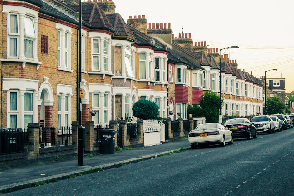 A row of traditional British terraced houses with brick facades and bay windows, each topped with chimney stacks, lining a residential street during daylight. Front gardens are enclosed with low brick walls and metal railings, some with small shrubs or potted plants. Cars are parked along the pavement, including a white vehicle closest to the camera, with other dark-colored cars further down the street. The pavement is clear, with a street lamp visible on the right side. In the foreground, a black wheelie bin is positioned near the curb. The surrounding environment appears calm, typical of a suburban area in Kennington, with a soft, warm light suggesting late afternoon or early evening. This scene exemplifies a typical UK residential street, relevant to home relocation and furniture transport services offered by Kennington Removals, especially when managing packing and loading processes for house moves in areas like SE11. Their professional team would handle the logistics of loading household furniture and boxes into vans for transportation.